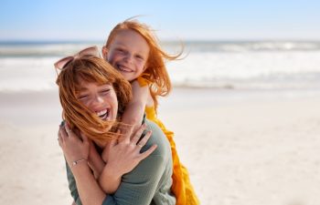 Smiling mother and beautiful daughter having fun at the beach. Portrait of happy woman giving a piggyback ride to cute little girl with copy space. Portrait of happy red hair kid embracing her mom.